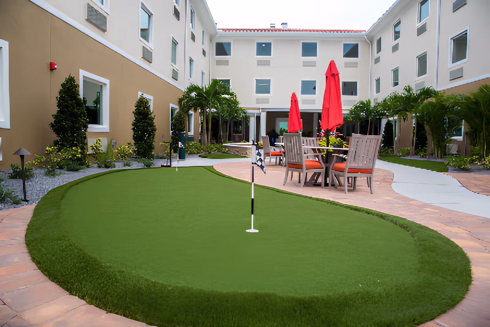 Outdoor courtyard area of a senior living facility with a small putting green, surrounded by a paved walkway. There are several chairs and tables with red umbrellas, and the courtyard is bordered by a three-story building with multiple windows and landscaped greenery.