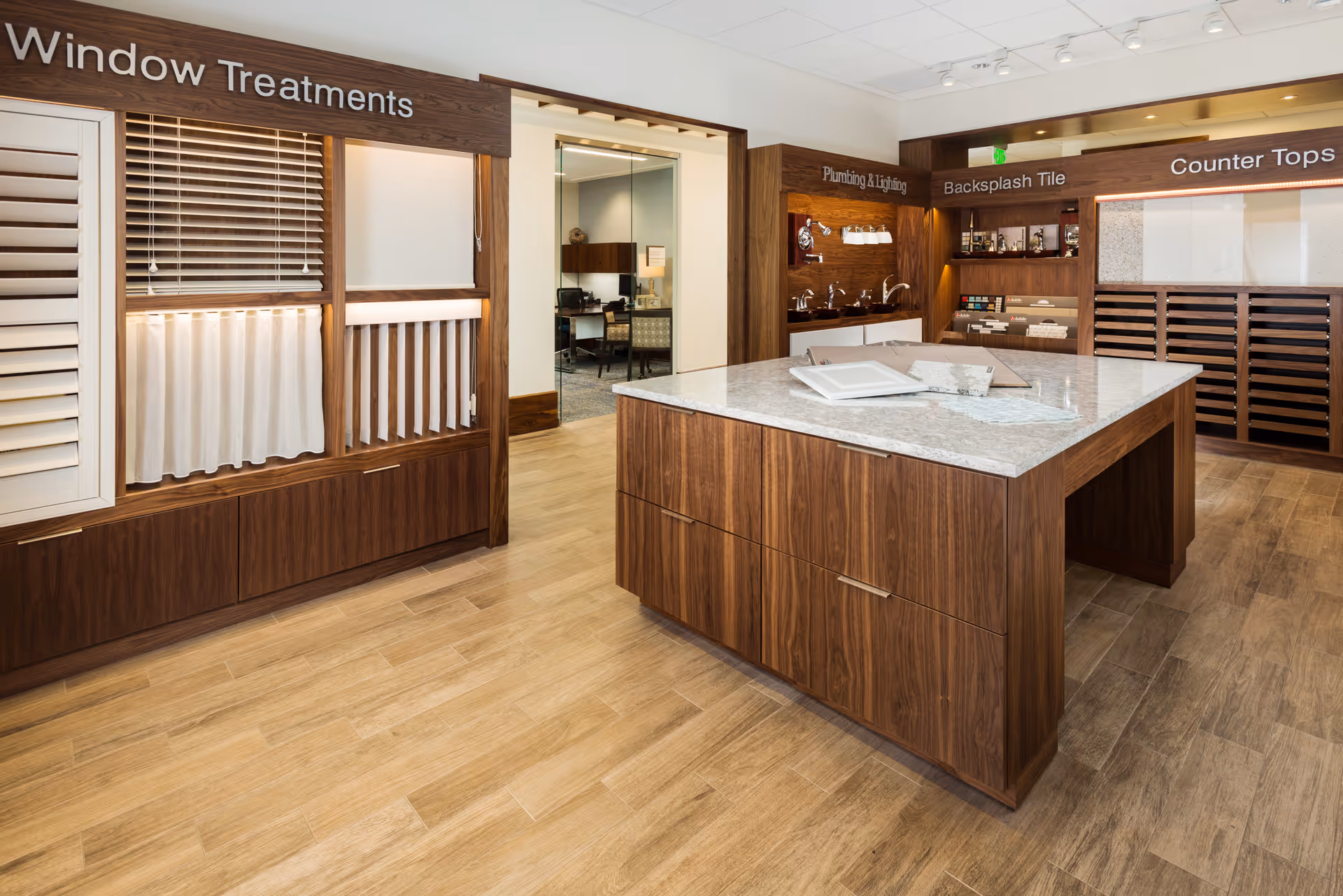Interior view of a showroom area in the Shell Point Welcome Center featuring displays of window treatments, plumbing and lighting fixtures, backsplash tile samples, and countertop materials. The room has wooden flooring, a large central island with a marble countertop, and various sample boards mounted on wooden panels.