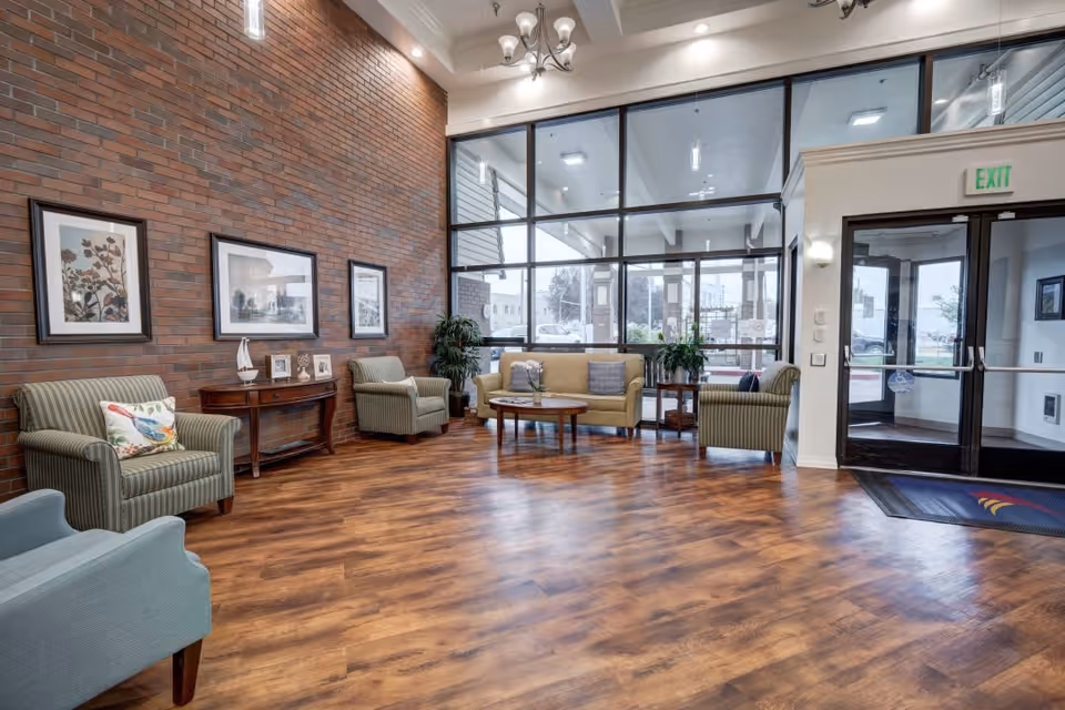 A spacious and well-lit senior living facility lobby with wooden flooring, a large brick accent wall adorned with framed artwork, several comfortable armchairs and a sofa arranged around a wooden coffee table, potted plants, and large glass windows and doors providing a view of the outside.