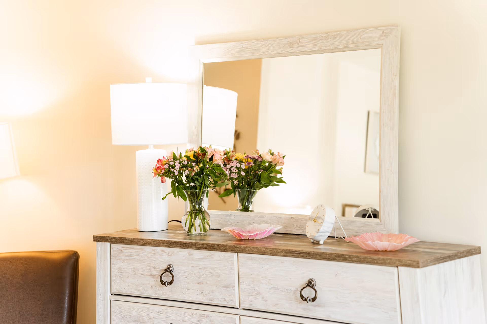 A wooden dresser with a large framed mirror on top. On the dresser are a white table lamp, a vase with colorful flowers, a small round clock, and two pink decorative dishes. Part of a brown chair is visible on the left side.