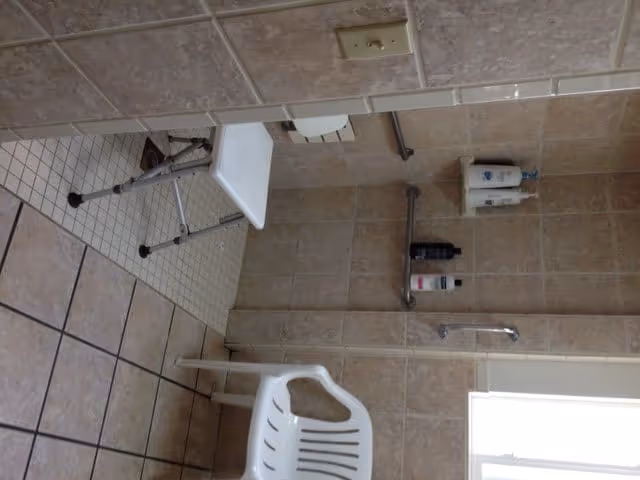 A tiled shower area with a white plastic chair and a white shower bench. There are grab bars on the walls and mounted bottles of shampoo and body wash. The floor has small square tiles in the shower and larger tiles outside the shower area.