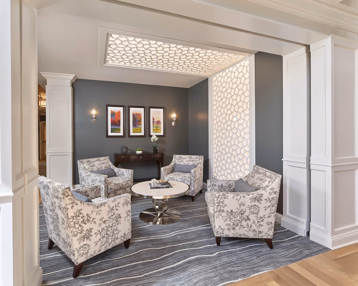 A cozy sitting area in an assisted living facility with four floral-patterned armchairs arranged around a round coffee table. The walls are painted gray with three framed landscape paintings and two wall sconces. A decorative illuminated panel is on one wall, and the floor is covered with a striped gray carpet.