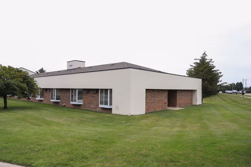 Exterior view of a single-story building with a combination of beige and brick walls, surrounded by a well-maintained green lawn. There are several windows along the side of the building and a tree on the left side. The sky is overcast.