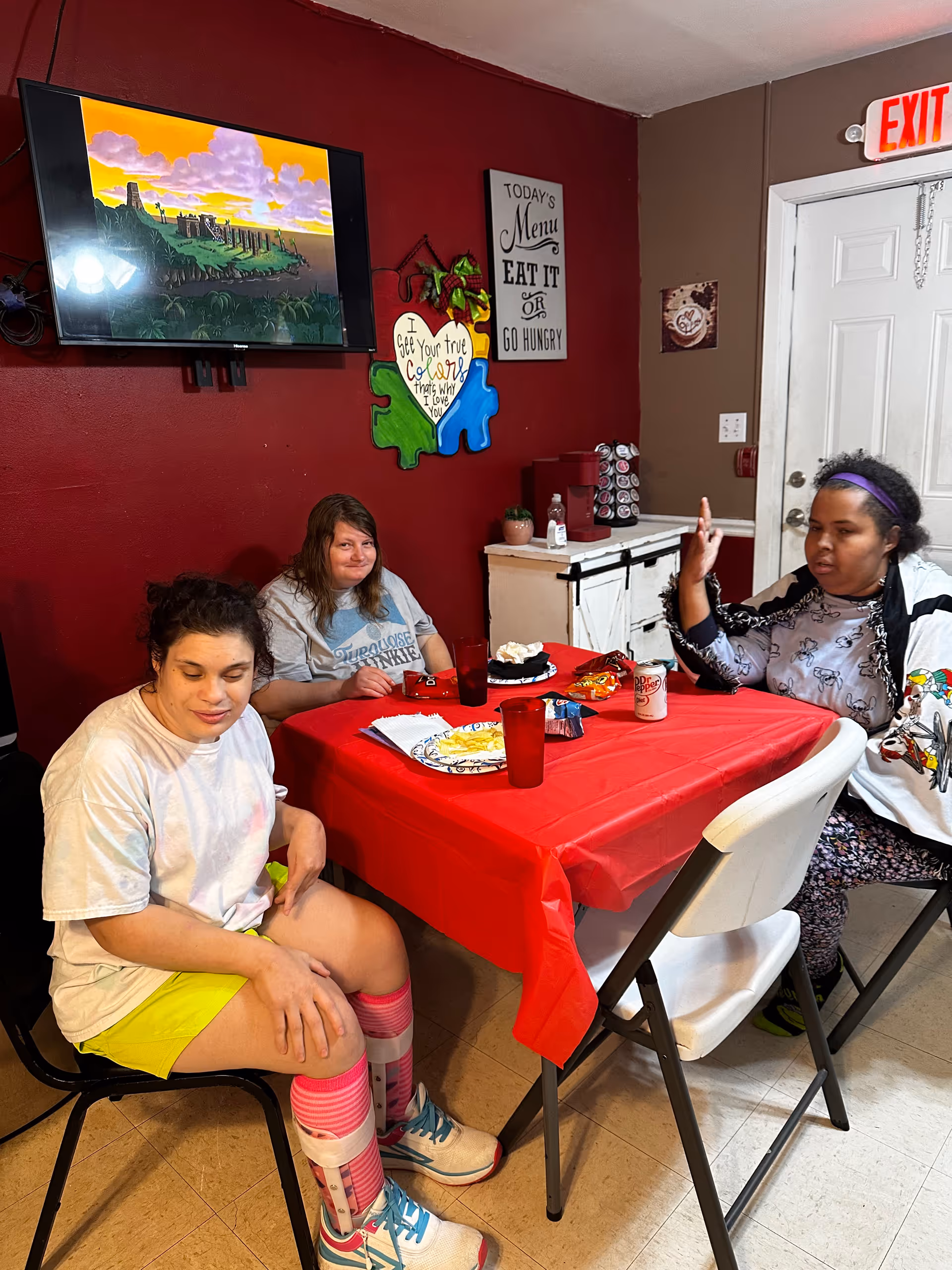 Three women sitting around a table with a red tablecloth in a room with red and brown walls. On the wall behind them is a TV displaying an animated scene, a colorful puzzle piece wall decoration with the text 'I See Your true Colors that's why I love you,' and a sign that says 'Today's Menu Eat It or Go Hungry.' The table has plates with food, drinks, and snacks. There is a white door with an exit sign above it in the background.