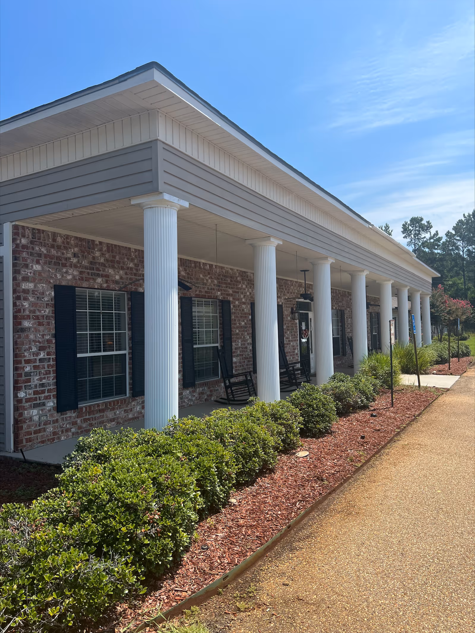 Front porch of a brick building with white columns, rocking chairs, manicured shrubs, and a paved walkway under a blue sky.