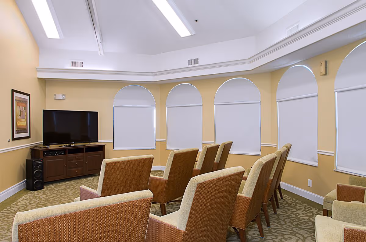 Small media room with rows of upholstered chairs facing a television beneath arched windows.