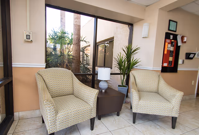 Two upholstered armchairs flank a small table with a lamp beside a window and potted plant in a facility lobby.