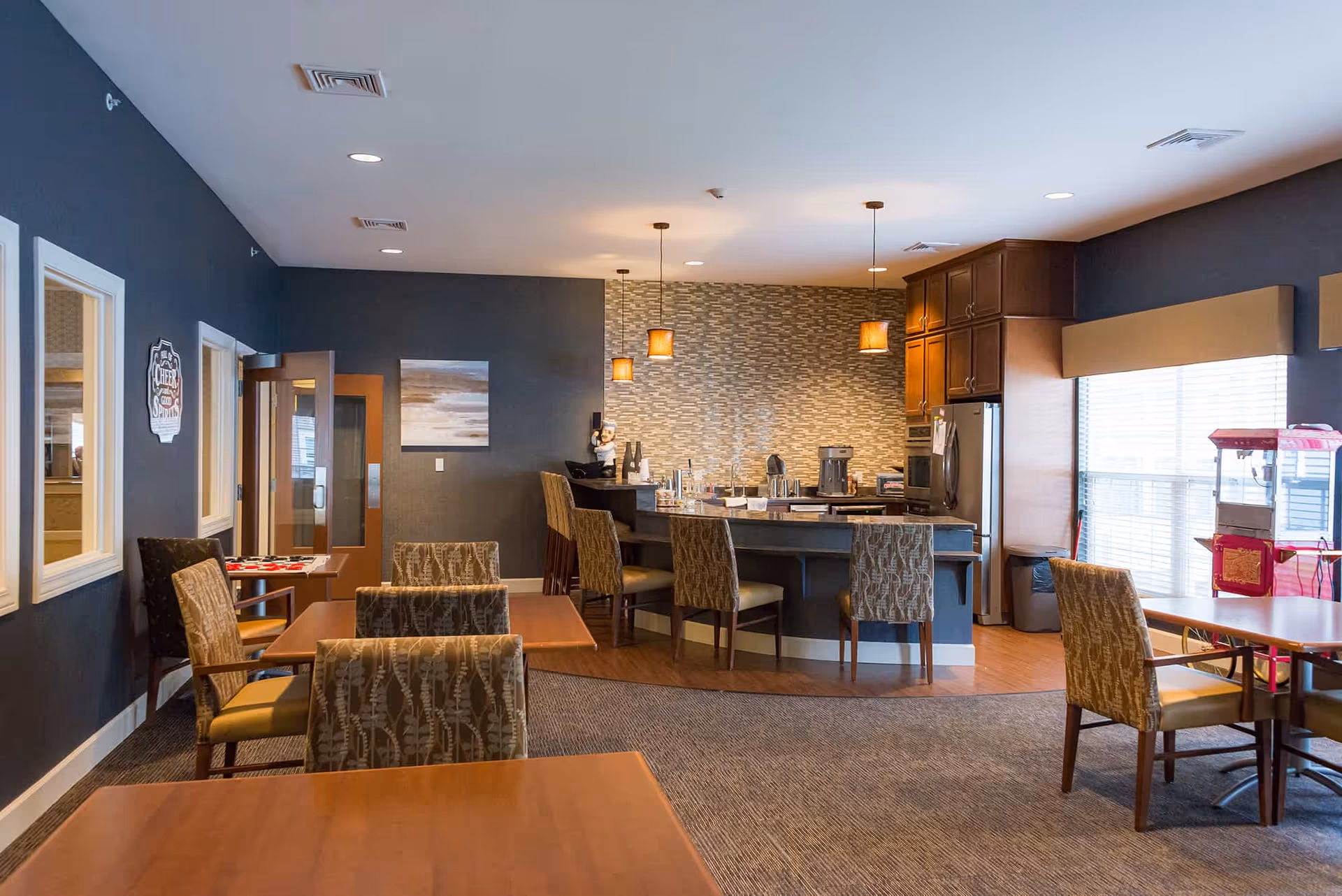 Communal dining area with tables and chairs facing a small kitchen/bar with pendant lights and large windows.