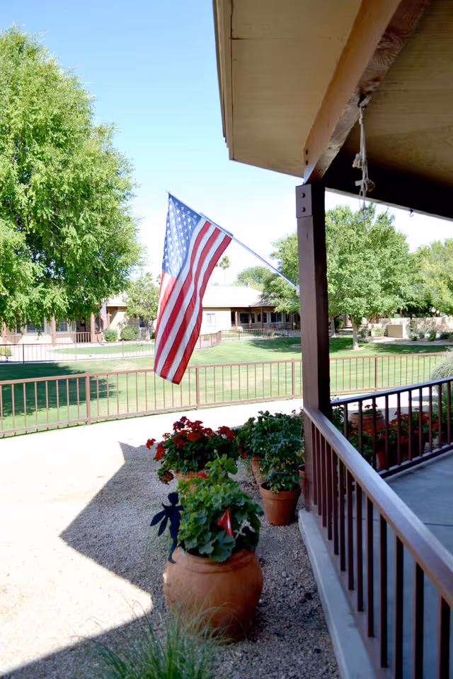 View from a covered porch with potted plants and an American flag hanging from a wooden post. In the background, there is a green lawn, trees, and a single-story building under a clear blue sky.