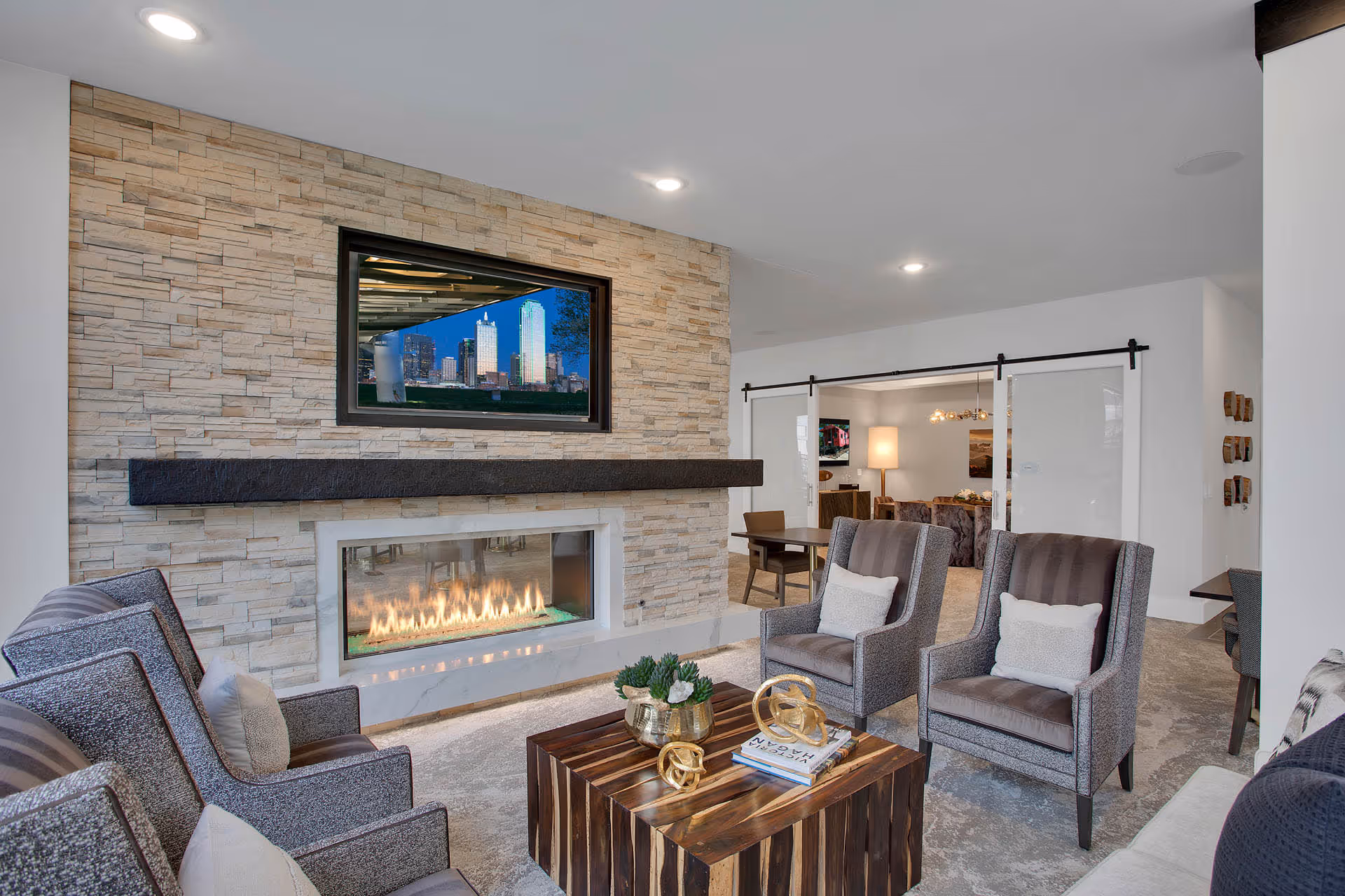 A modern living room with a stone accent wall featuring a built-in gas fireplace and a flat-screen TV mounted above it. The room has four upholstered armchairs with pillows arranged around a wooden coffee table decorated with books and small sculptures. In the background, sliding barn doors open to another room with a lamp and additional seating.