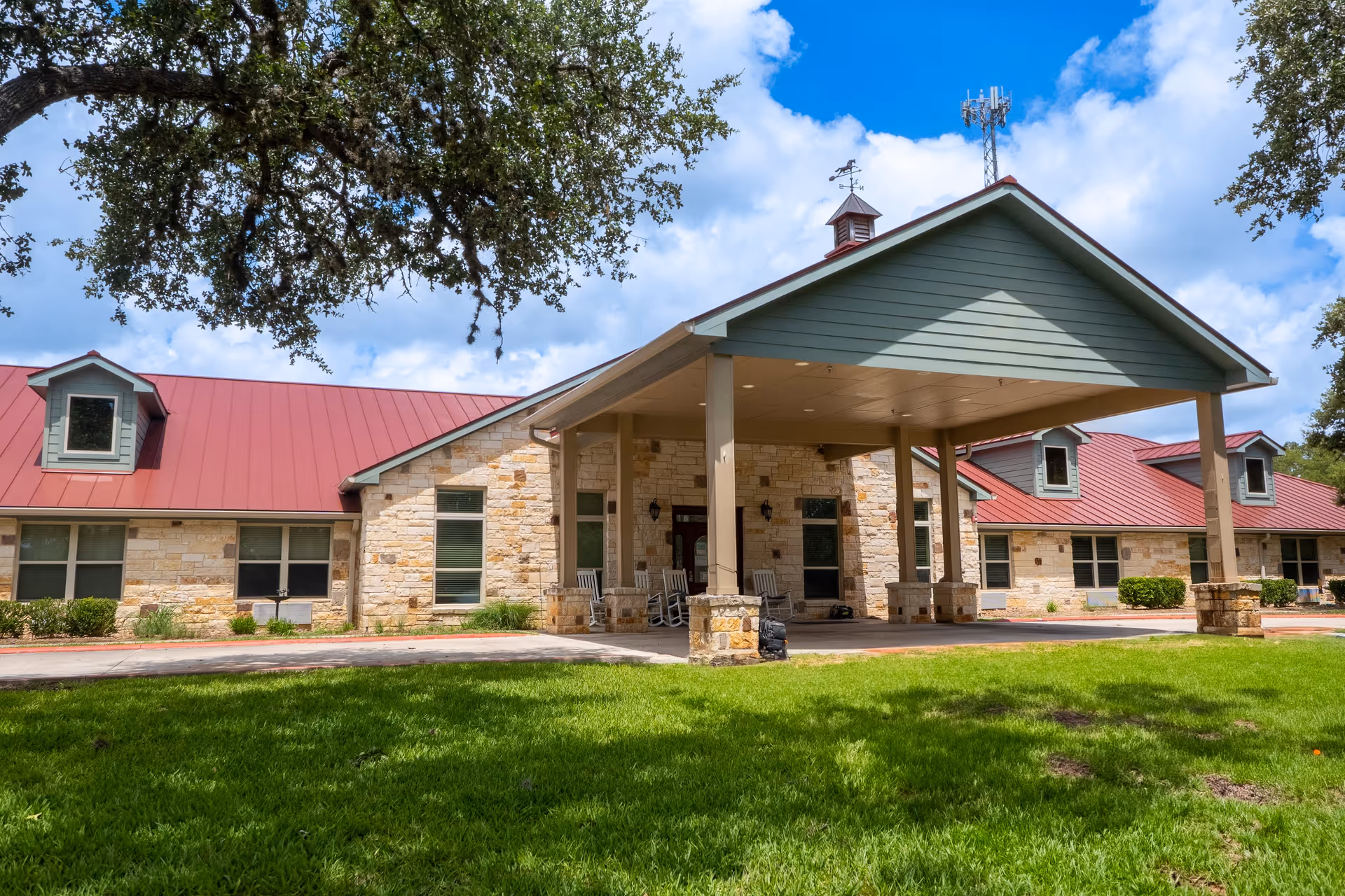 Exterior view of Gemstone Senior Living at Bulverde, showing a single-story building with stone walls, a red metal roof, and a covered entrance supported by columns. There is a well-maintained green lawn in front and a large tree providing shade.