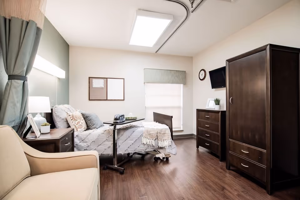 A well-lit nursing facility bedroom with a hospital-style bed covered in a white and gray patterned blanket and pillows. Next to the bed is a small wooden nightstand with a lamp and a small plant. There is a beige armchair in the foreground, a wooden dresser with a small plant and picture frame, a wall-mounted TV, a clock, and a large wooden wardrobe. The room has wood flooring, a window with a blind and valance, and a bulletin board on the wall.
