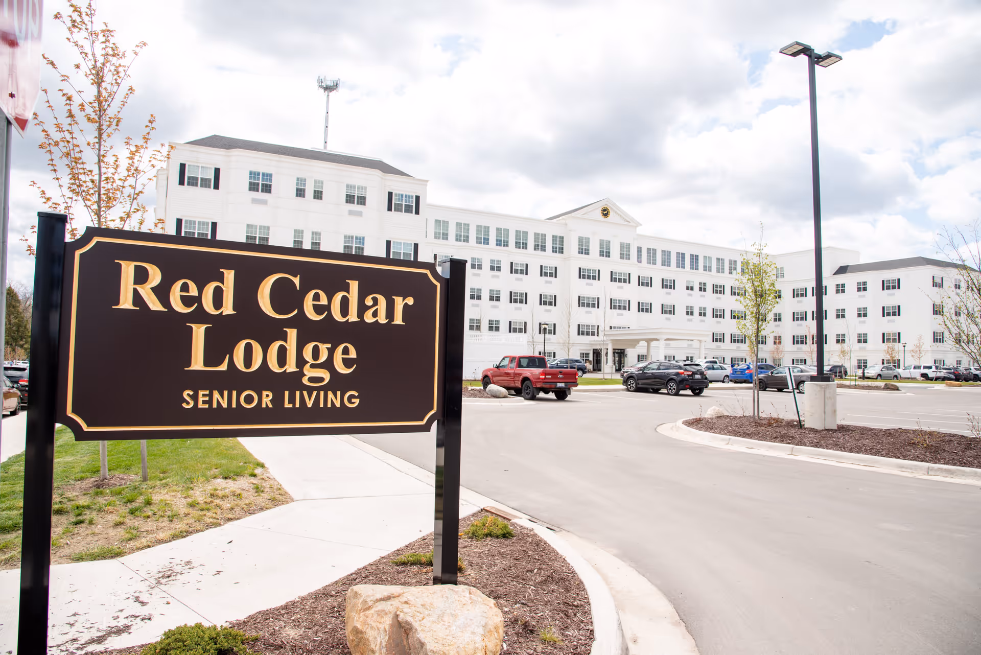 Exterior view of Red Cedar Lodge senior living facility with a large white multi-story building, parking lot with several cars, and a prominent sign in the foreground displaying the facility's name.