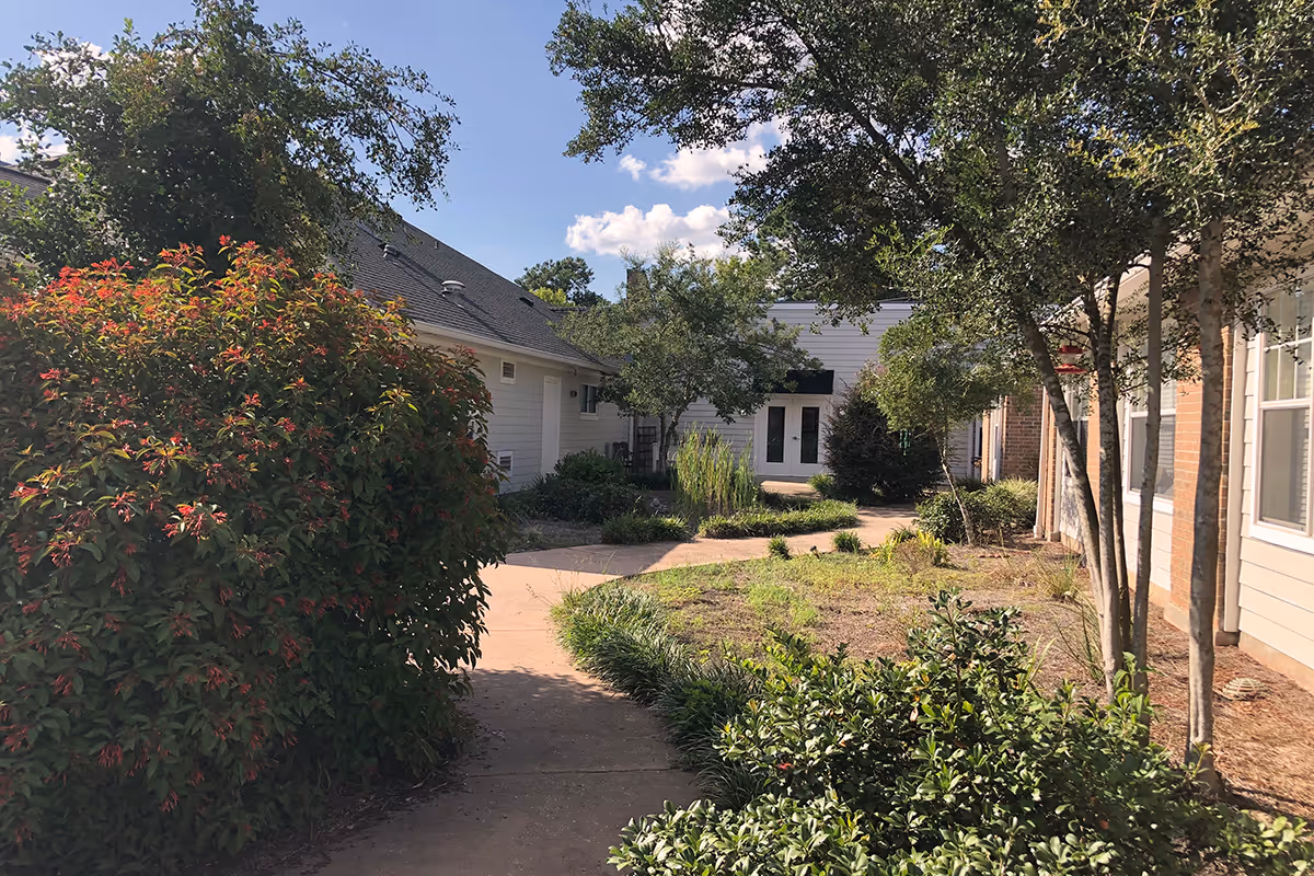 Outdoor courtyard area at Oak Park Village at Slidell featuring a paved walkway surrounded by green bushes, trees, and plants with buildings on either side under a blue sky with some clouds.