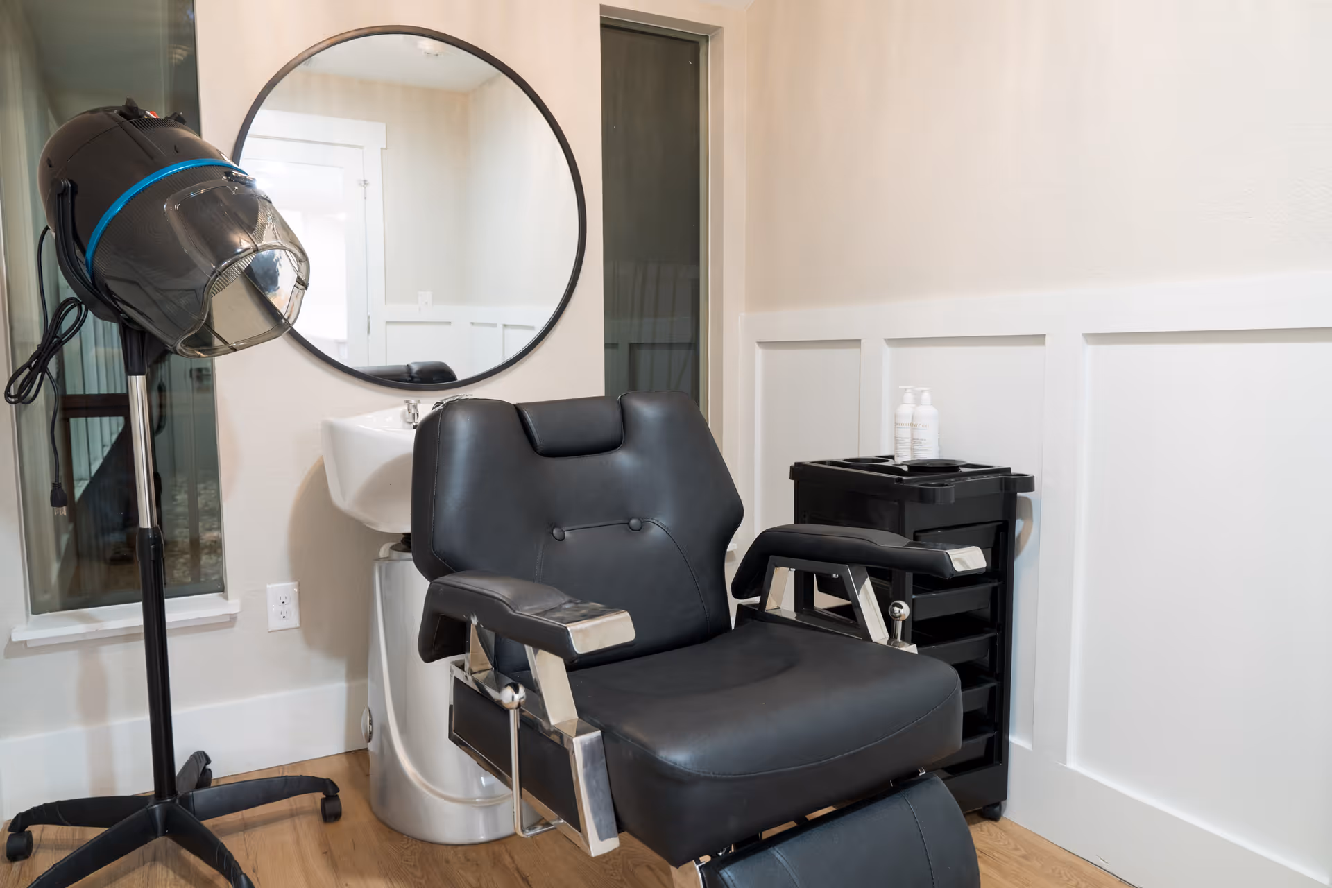 A small salon area with a black salon chair, a hair dryer on a stand, a round mirror mounted on the wall, a white sink, and a black rolling cart with hair products in a room with light-colored walls and wooden flooring.
