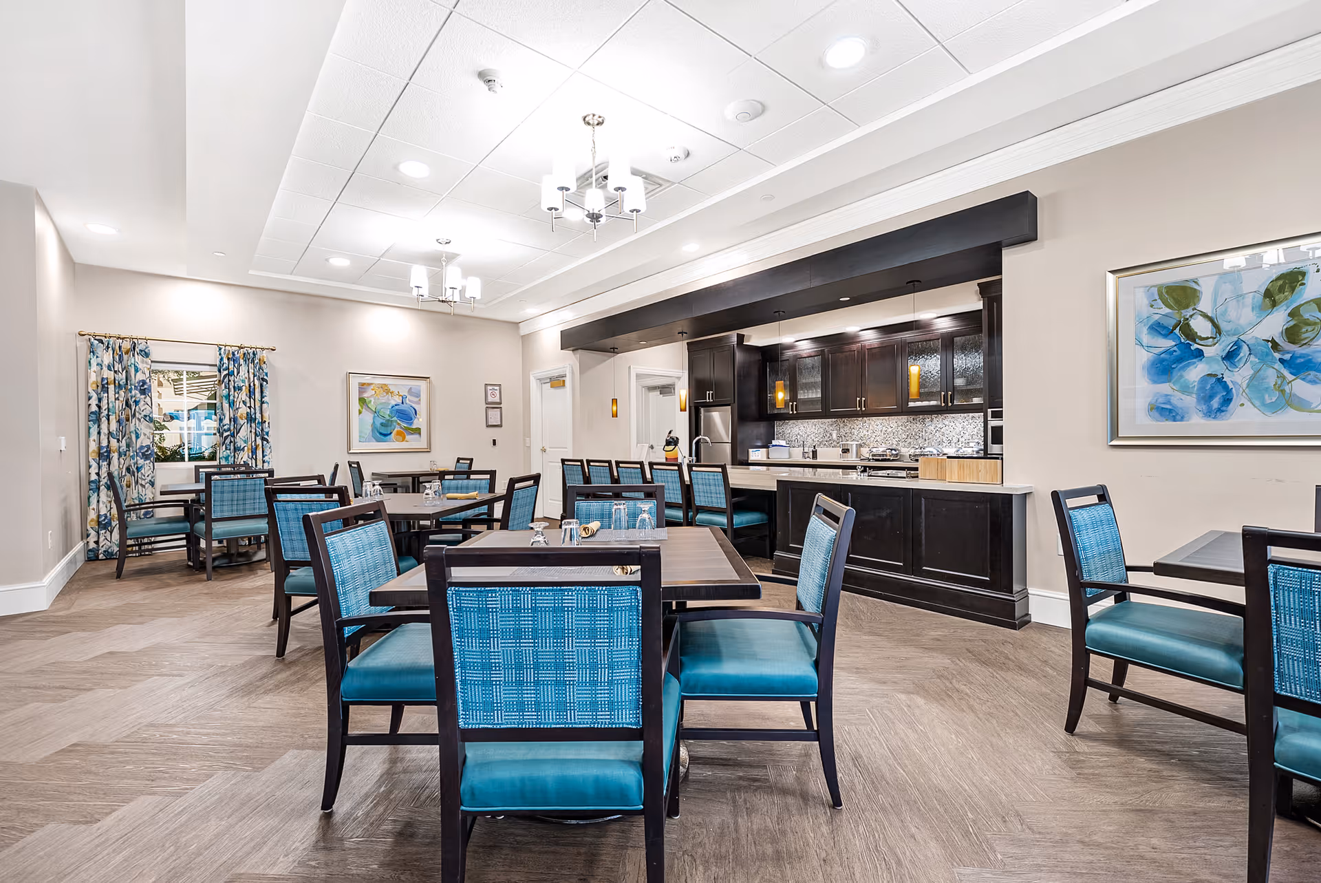 A bright dining room with multiple tables and chairs featuring blue upholstery. The room has light-colored walls, decorative curtains, framed artwork, and a modern kitchen area with dark cabinetry and stainless steel appliances in the background.