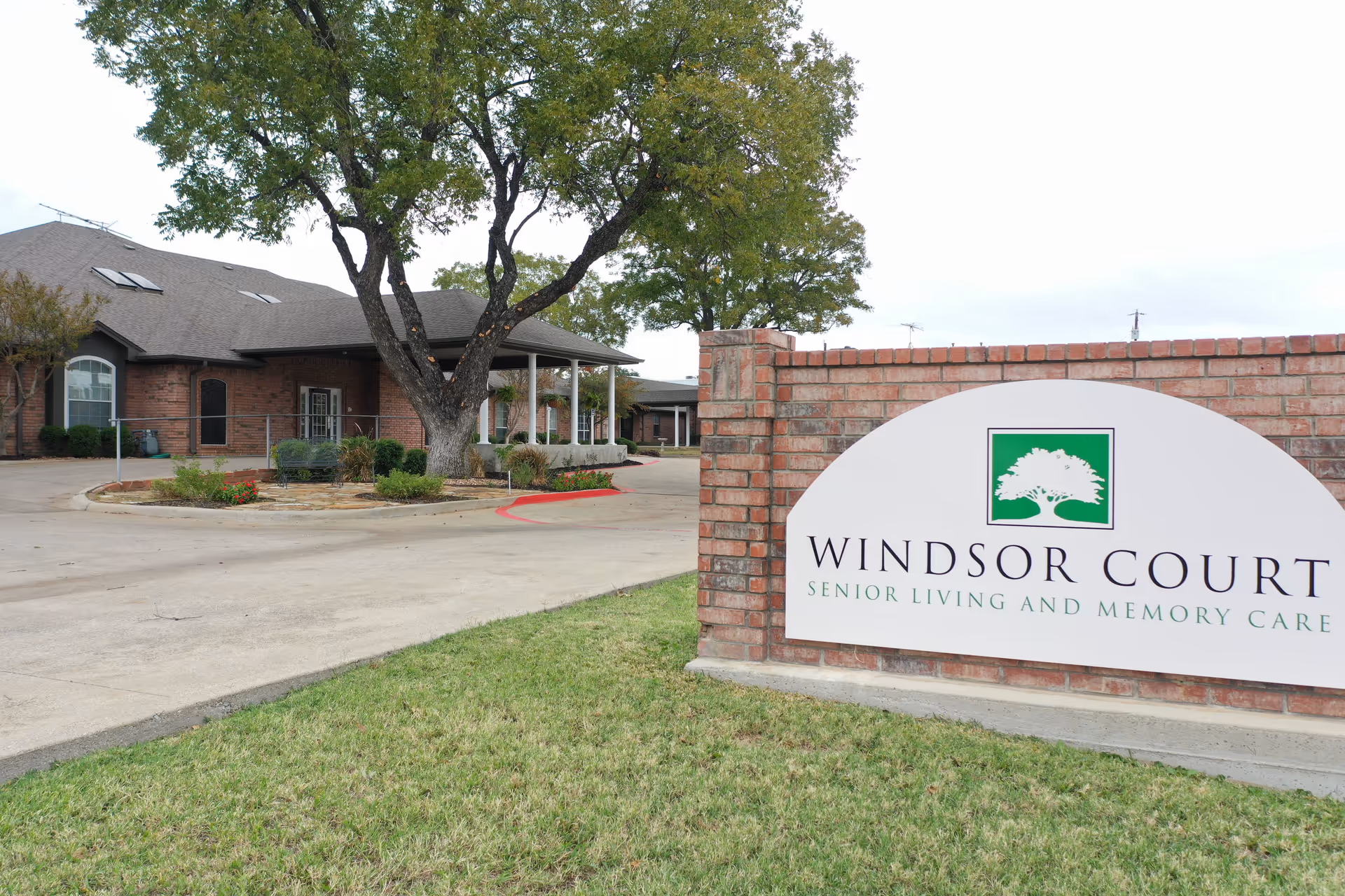 Exterior view of Windsor Court Senior Living and Memory Care facility showing a brick building with a covered entrance, a large tree, and a brick sign with the facility's name and logo on a grassy lawn.