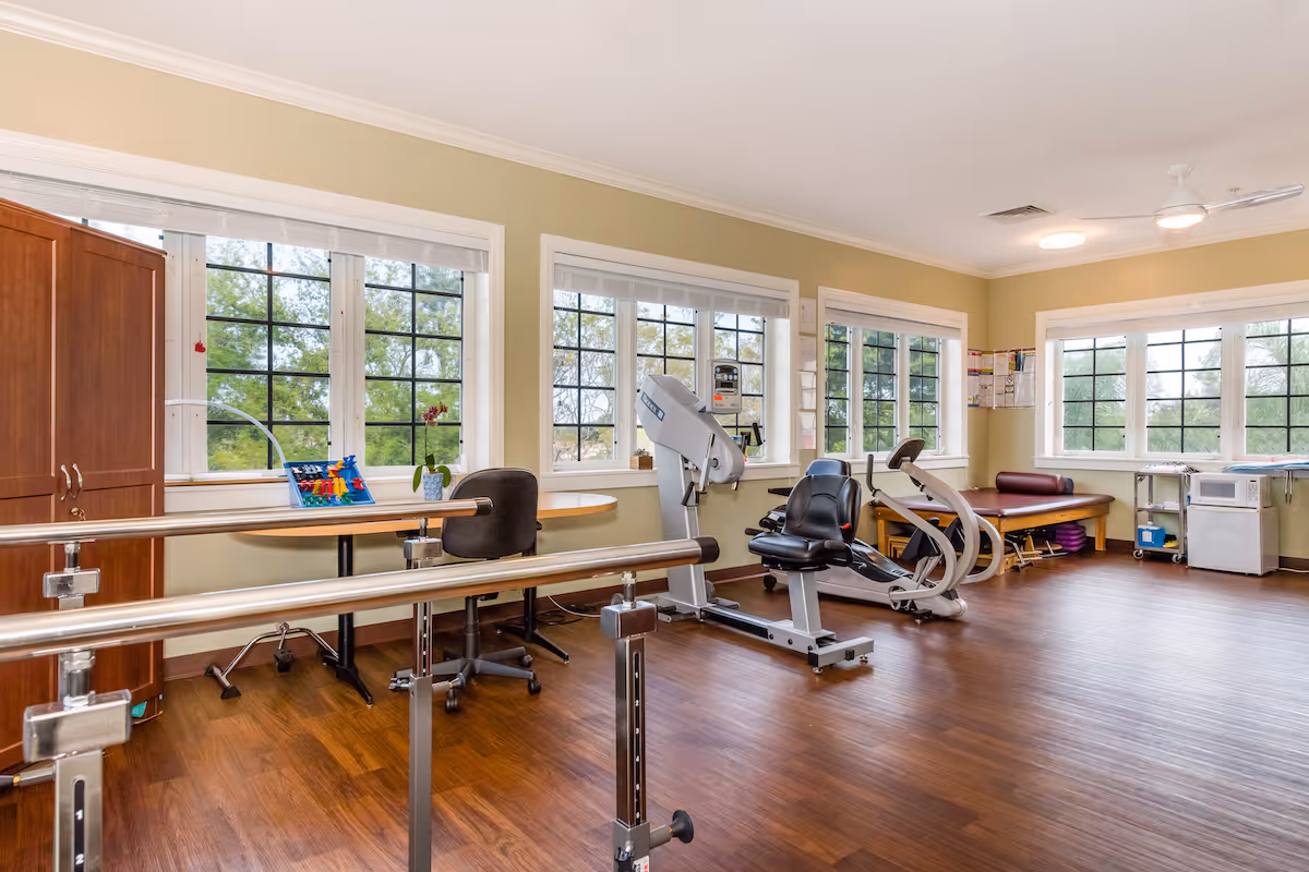 Bright therapy/exercise room with recumbent exercise machines, parallel bars, chairs and large windows.