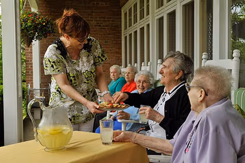 A caregiver serving snacks to a group of elderly women seated on a covered porch with rocking chairs. A table with a pitcher of lemonade and glasses is in the foreground.