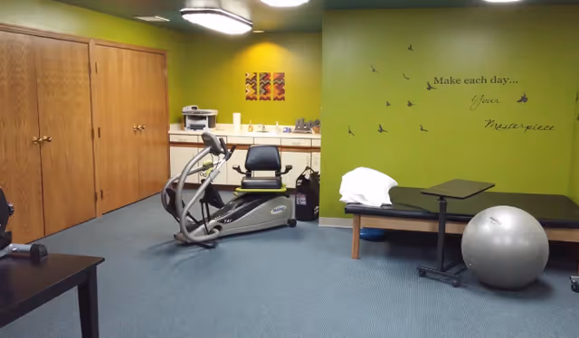 A senior living facility room with green walls and blue carpet featuring exercise equipment including a recumbent bike and a large exercise ball. There is a bench with a pillow and a small adjustable table. On the wall, there is a motivational quote that reads 'Make each day... Your Masterpiece' with bird decals flying around it. Wooden cabinets and a countertop with a printer and other items are visible in the background.