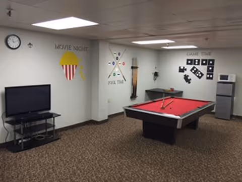Interior recreation room with a red pool table, television on a stand, wall game-themed decals, and a small refrigerator.