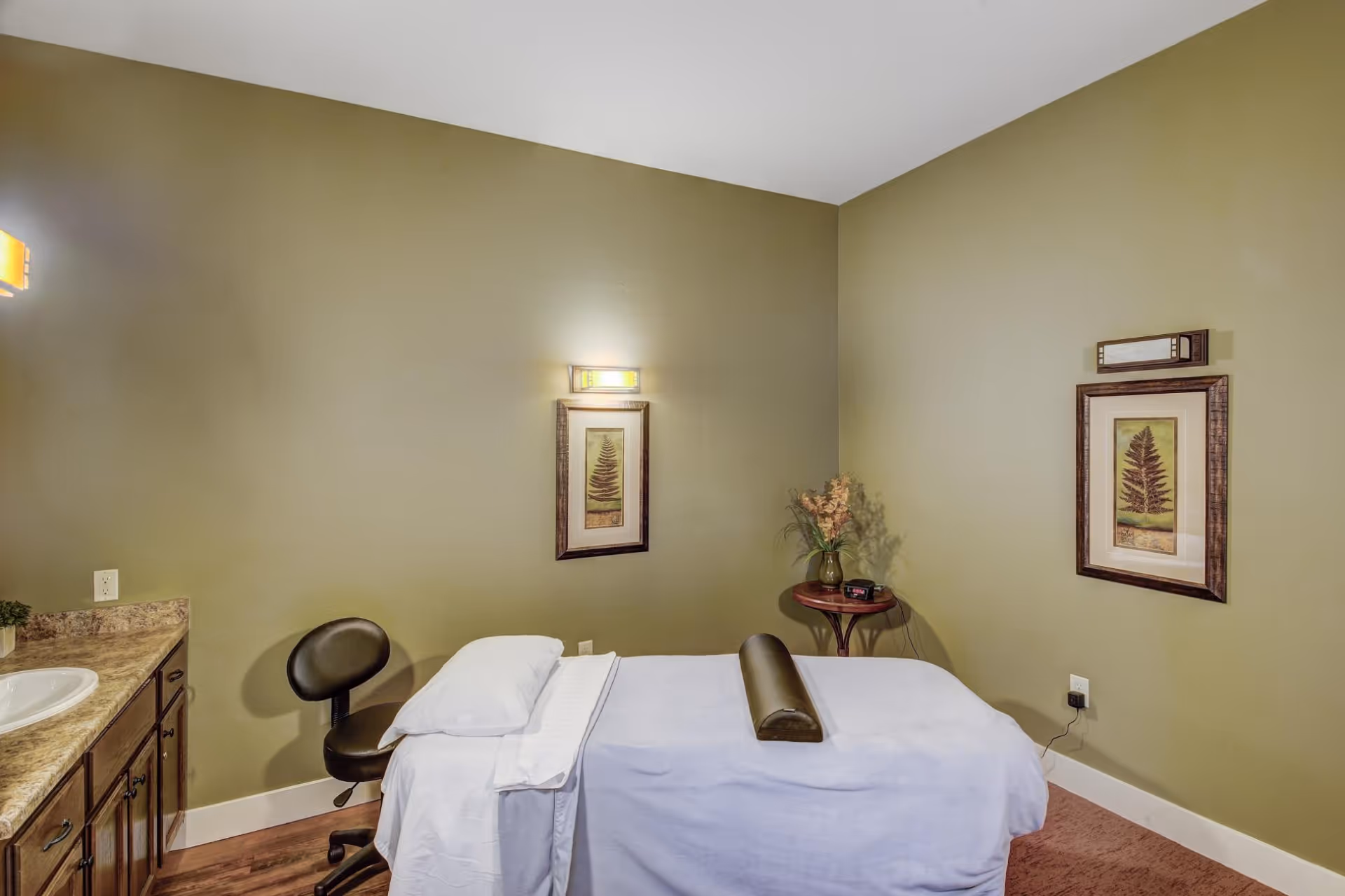 Small treatment room with a massage table, rolling stool, sink vanity and framed artwork against olive-green walls.