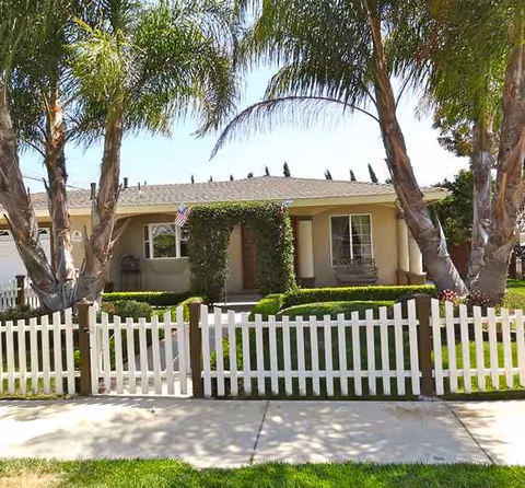 A single-story beige building with a white picket fence and palm trees in front.