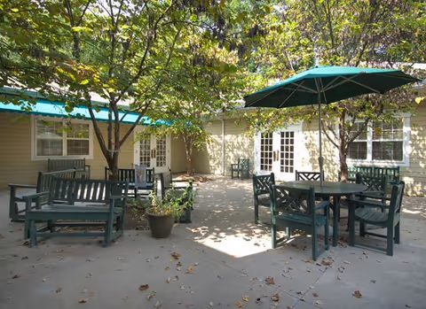 Outdoor patio area with green wooden benches and tables, some shaded by large green umbrellas, surrounded by trees and adjacent to a light-colored building with multiple windows and doors.