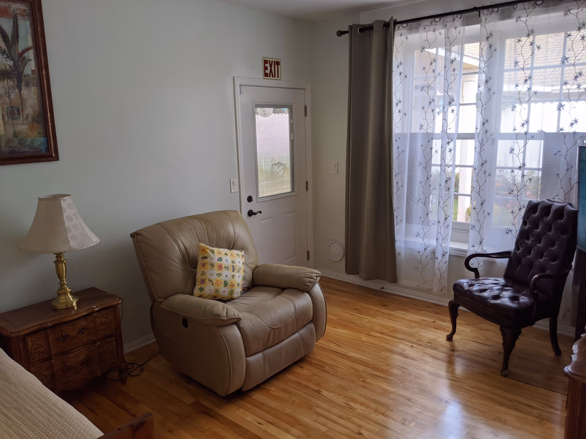 A cozy living room area with a beige leather recliner chair featuring a decorative pillow, a wooden side table with a brass lamp, a dark brown tufted armchair, a window with sheer floral curtains, and a white door with an exit sign above it. The room has light-colored walls and hardwood flooring.