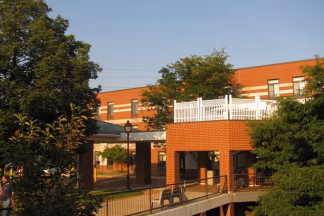 Exterior view of a red brick building with white trim, surrounded by trees and greenery. The building has a covered walkway and a raised patio area with white fencing. The scene is lit by warm sunlight.