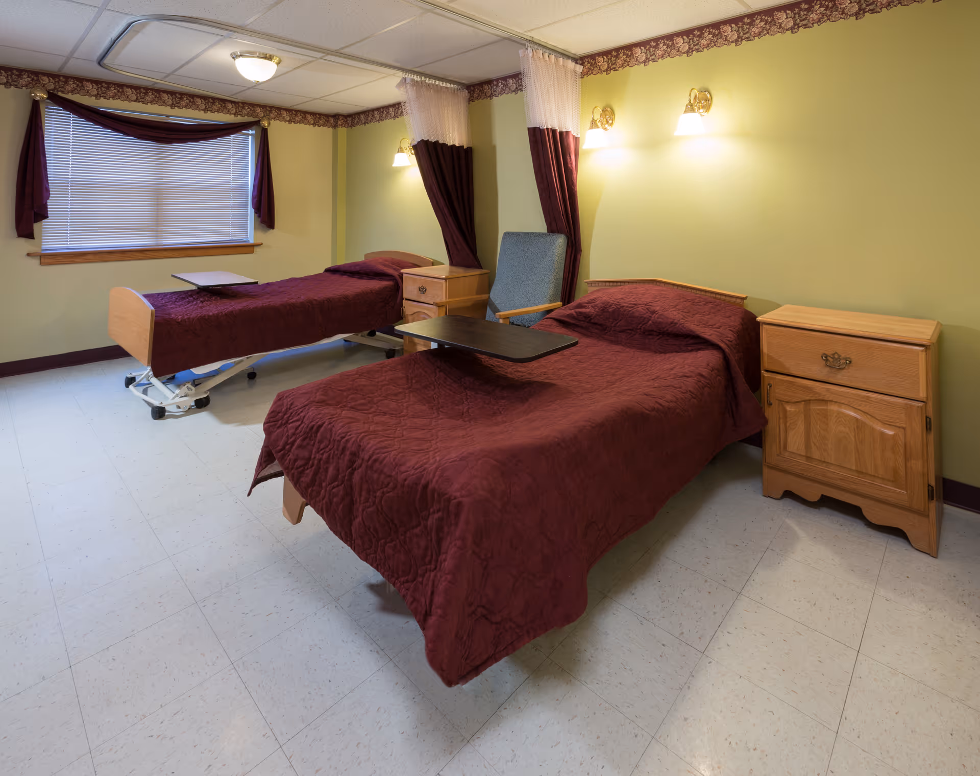 A room with two single beds covered in maroon bedspreads, each with an overbed table. There are two wooden nightstands, a blue upholstered chair, yellow-green walls with floral wallpaper border near the ceiling, and a window with blinds and maroon curtains.