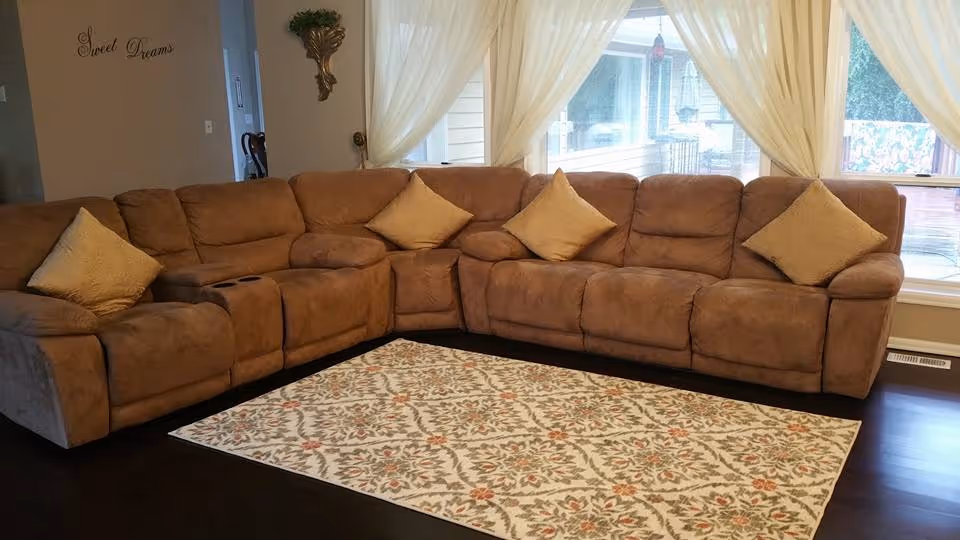 A cozy living room with a large brown sectional sofa adorned with four beige throw pillows. The sofa is positioned on a decorative area rug with a floral pattern. Behind the sofa, large windows with sheer white curtains allow natural light to fill the room. On the wall to the left, the words 'Sweet Dreams' are displayed in cursive lettering.