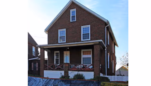 A two-story brick house with a covered front porch and several windows, set against a blue sky.