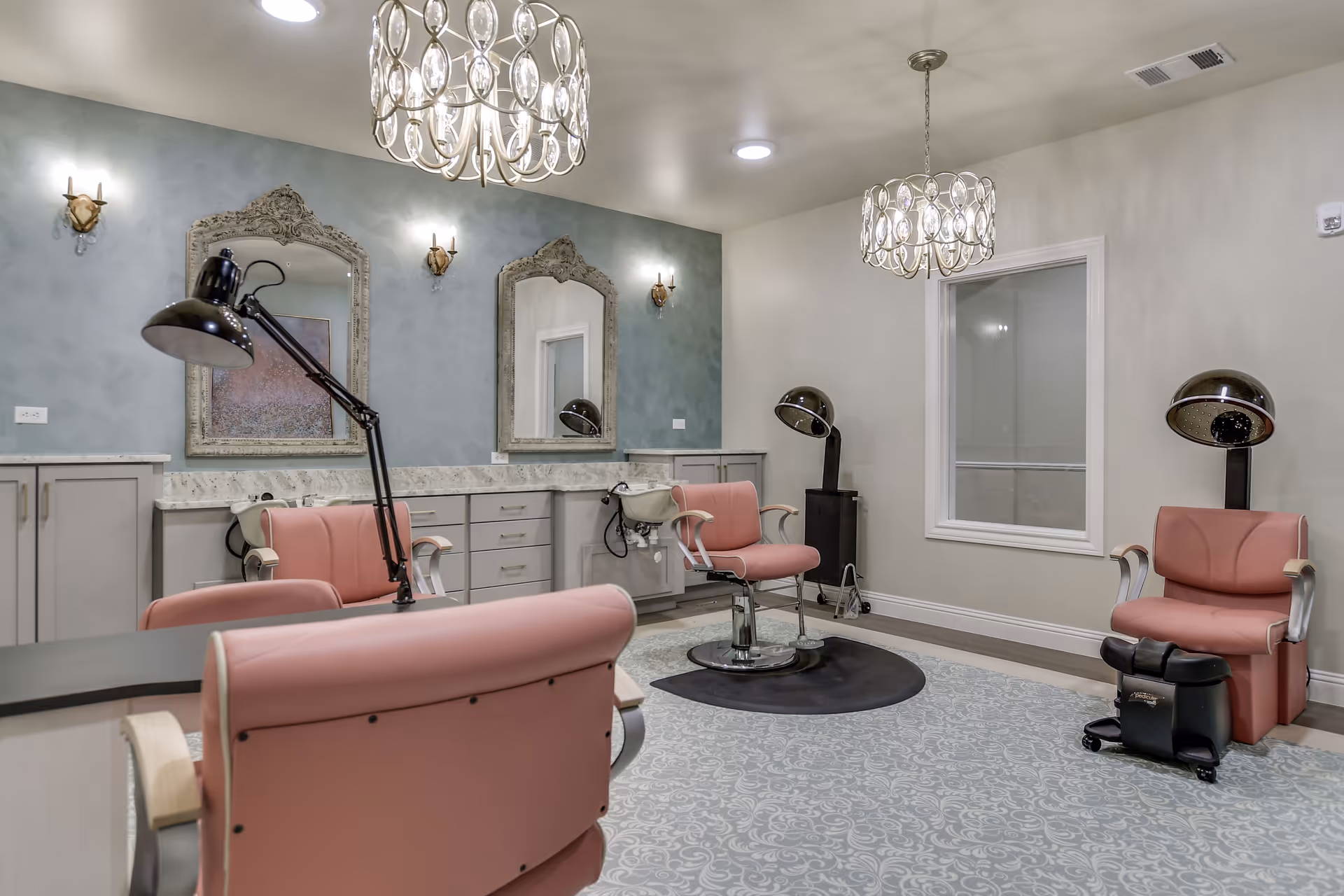 Interior of a salon room with pink salon chairs, two ornate mirrors on a blue accent wall, hair drying stations, and modern chandeliers hanging from the ceiling.