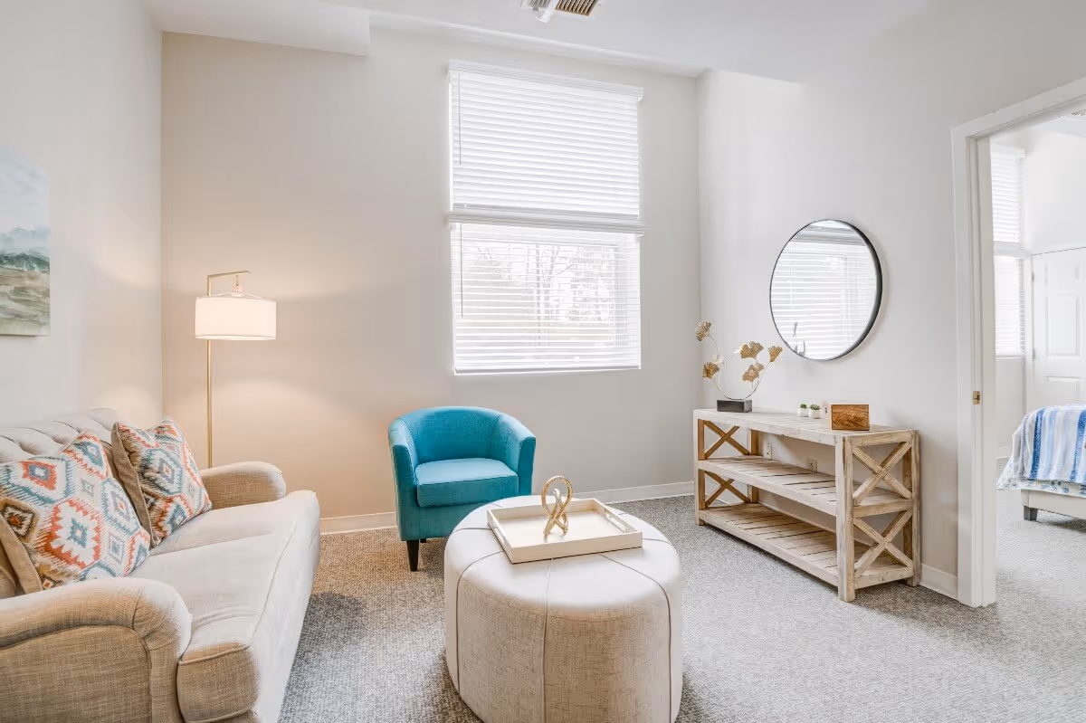 Bright furnished living room with a sofa, patterned pillows, teal armchair, round ottoman, console table and a window with blinds.