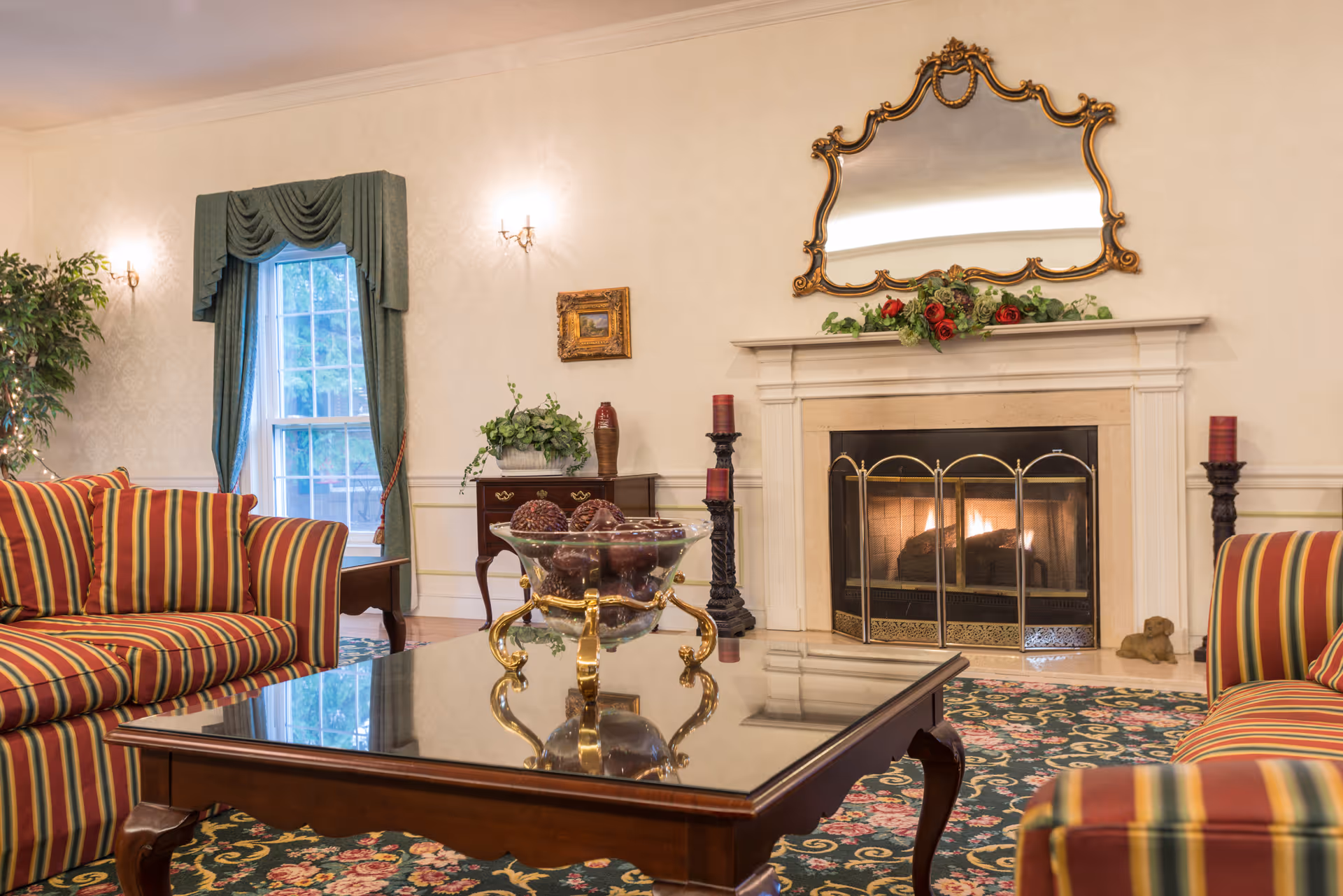 A cozy living room with a lit fireplace, two striped sofas facing each other, a glass-top wooden coffee table with a decorative bowl, a large ornate mirror above the fireplace, green curtains framing a window, and a floral patterned carpet.