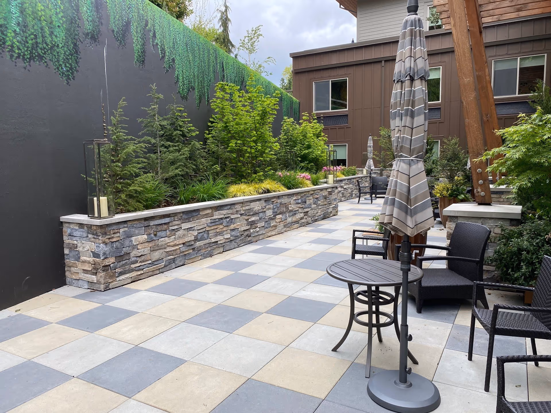 Outdoor patio area with checkered tile flooring, stone planter filled with green plants and small trees, several black wicker chairs, and round tables with closed striped umbrellas. The area is adjacent to a building with brown siding and windows.