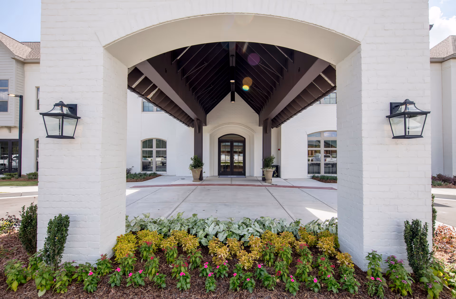 Entrance of The Blake at Woodcreek Farms featuring a covered driveway with a dark wooden roof structure, white brick pillars, wall-mounted lantern-style lights, and a landscaped flower bed with green and yellow foliage and pink flowers in front.