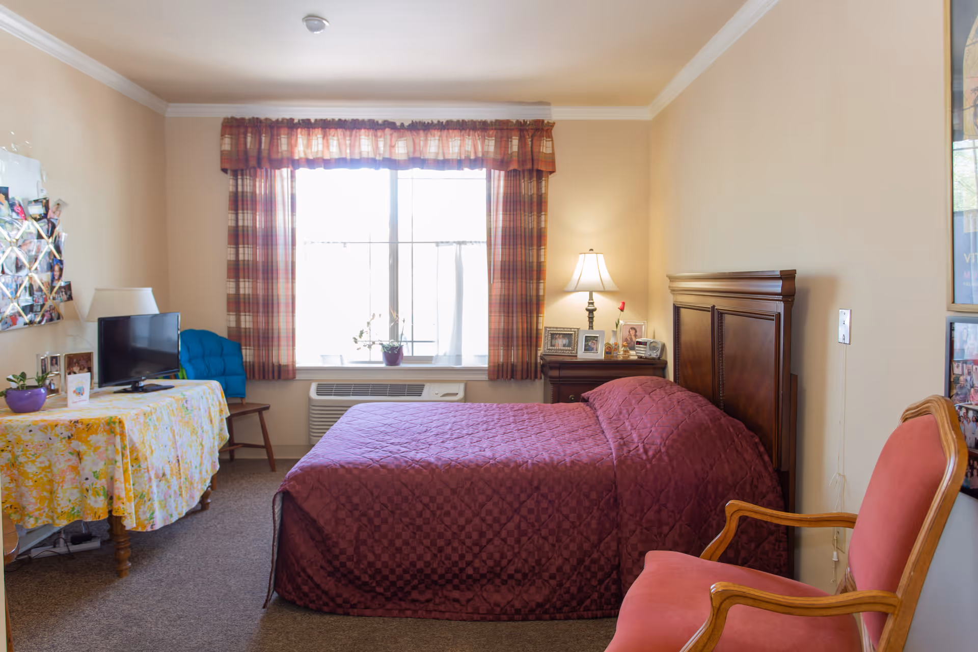 A cozy bedroom in an assisted living facility with a bed covered in a maroon quilt, a wooden headboard, a nightstand with a lamp and framed photos, a window with plaid curtains, a table covered with a floral tablecloth holding a small TV and a purple potted plant, and a red upholstered chair with wooden arms.