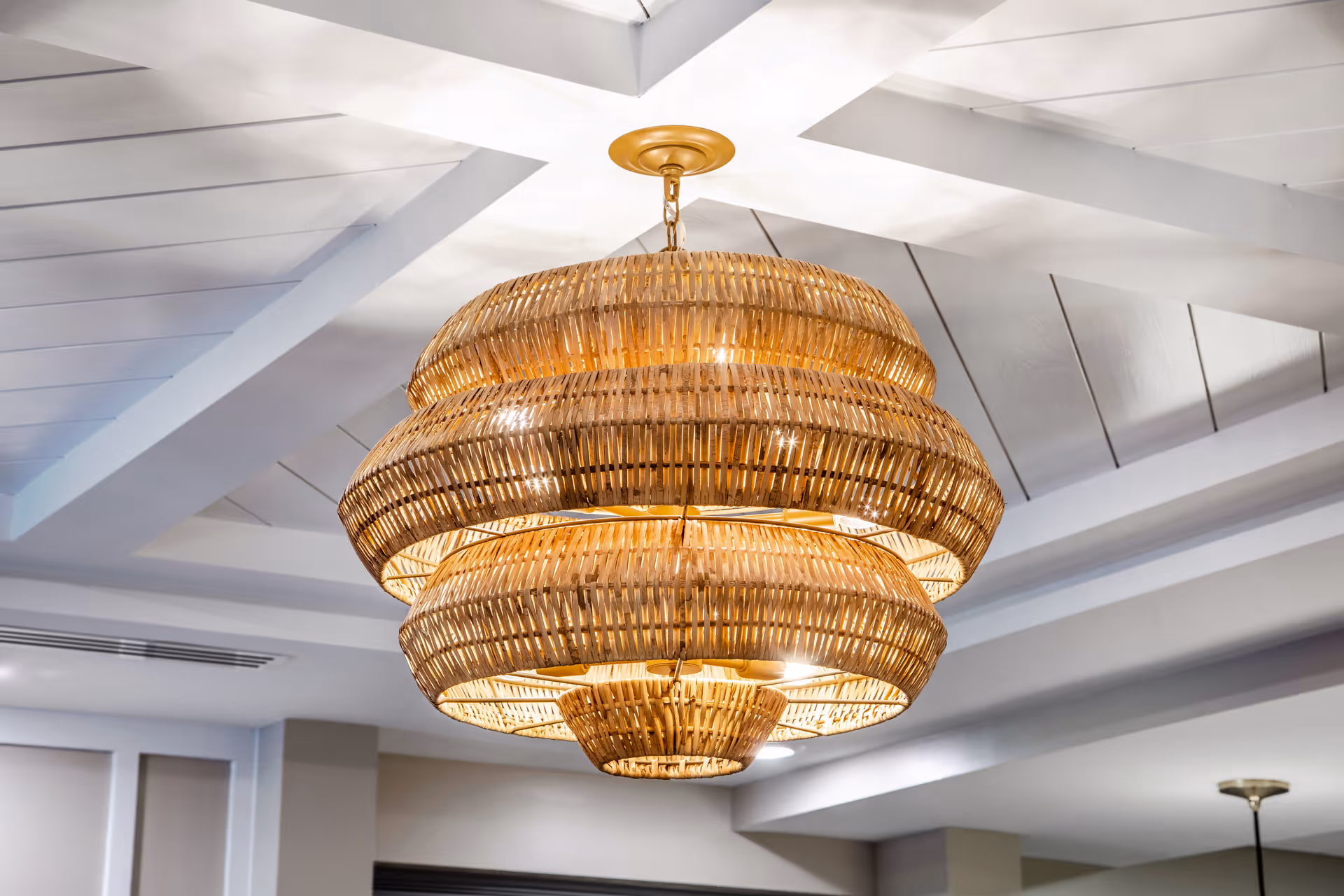 A large, multi-tiered woven rattan chandelier hanging from a white ceiling with exposed beams in an interior space.