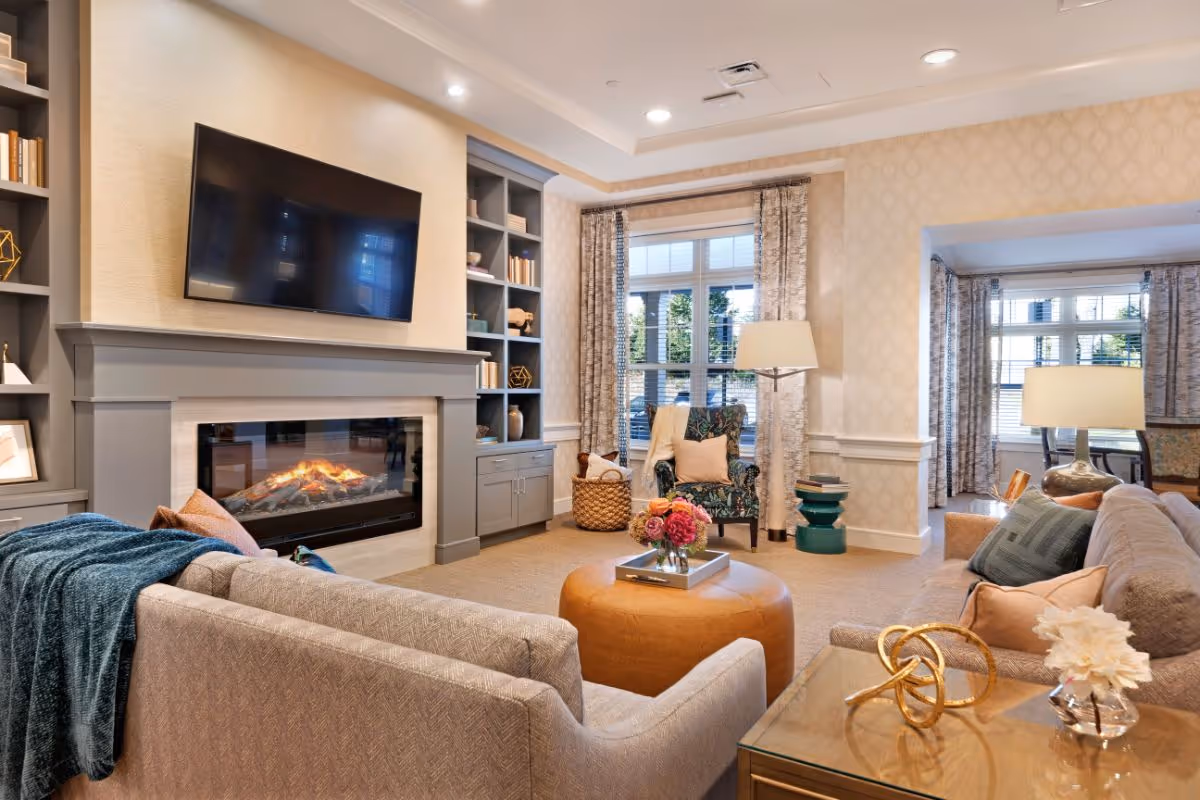 A cozy living room with a modern fireplace beneath a mounted flat-screen TV. The room features built-in shelves with books and decorative items, a large window with patterned curtains, a floral armchair, a round leather ottoman with a flower vase on top, and comfortable sofas with cushions and a throw blanket. There are also two floor lamps and a side table with decorative objects.