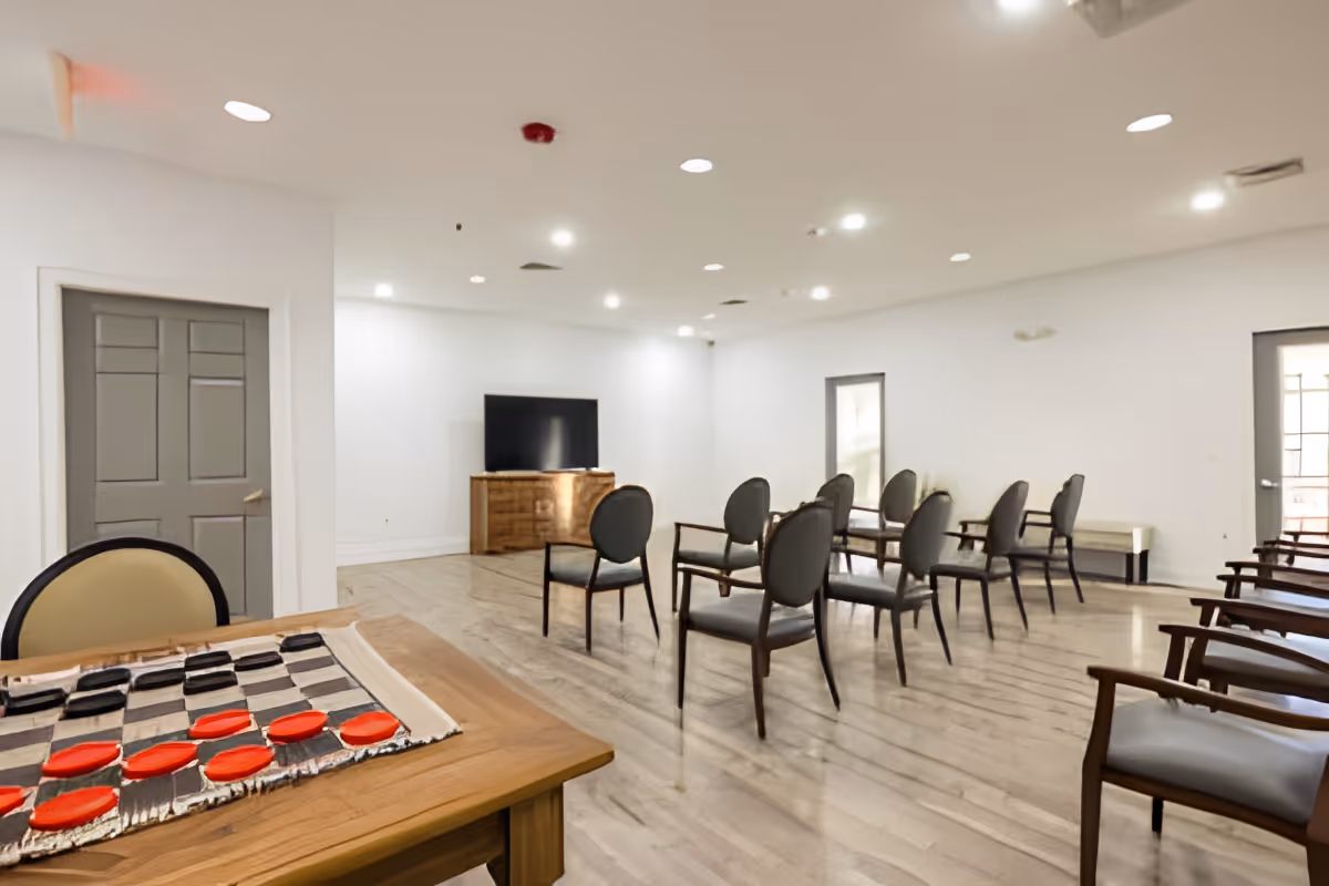 A spacious room with multiple chairs arranged in rows facing a flat-screen TV on a wooden cabinet. In the foreground, there is a table with a checkers board and pieces set up. The room has white walls, wooden flooring, recessed ceiling lights, and two gray doors.