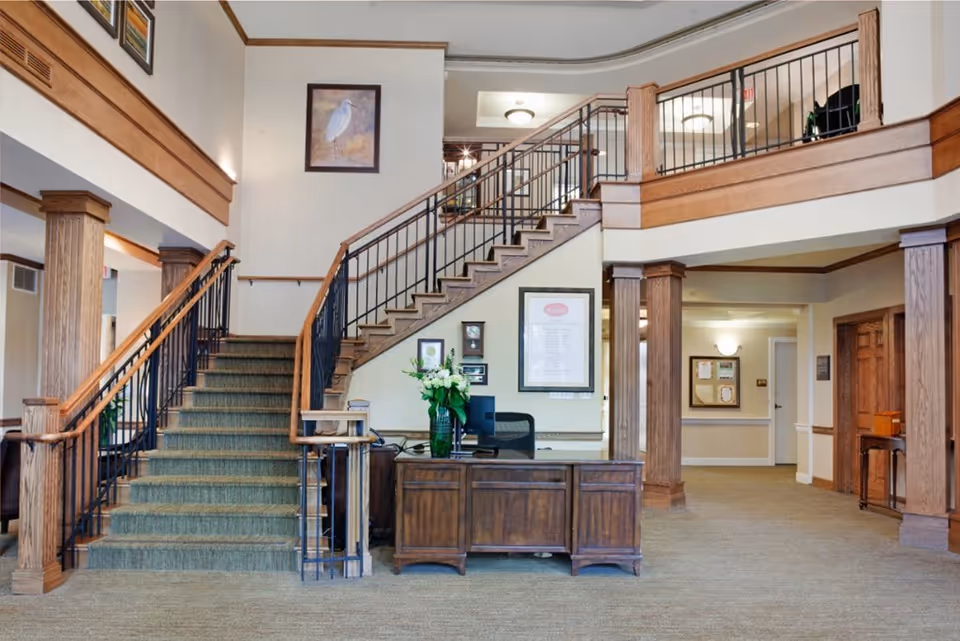 Interior view of a senior living facility lobby featuring a central wooden staircase with black metal railings, a wooden reception desk with a vase of flowers, framed artwork on the walls, and a carpeted floor. The space is well-lit with ceiling lights and has wooden columns and trim.