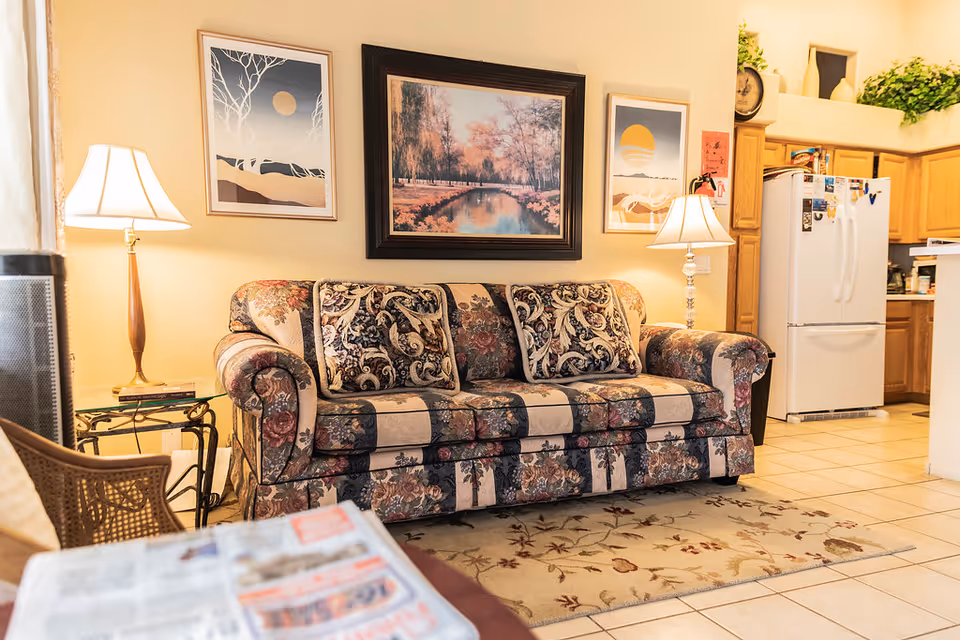 A cozy living room area with a floral patterned sofa adorned with three matching cushions. Two table lamps with white shades are placed on either side of the sofa on small tables. Above the sofa, there are three framed artworks hanging on the wall. To the right, a white refrigerator and wooden kitchen cabinets are visible, indicating an open layout connecting the living room and kitchen. The floor is tiled, and a floral area rug is placed in front of the sofa.