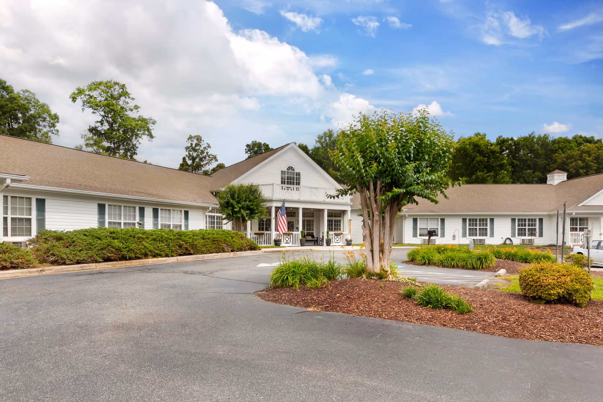 Front exterior of a single-story white senior living building with a circular driveway, landscaping, and an American flag.