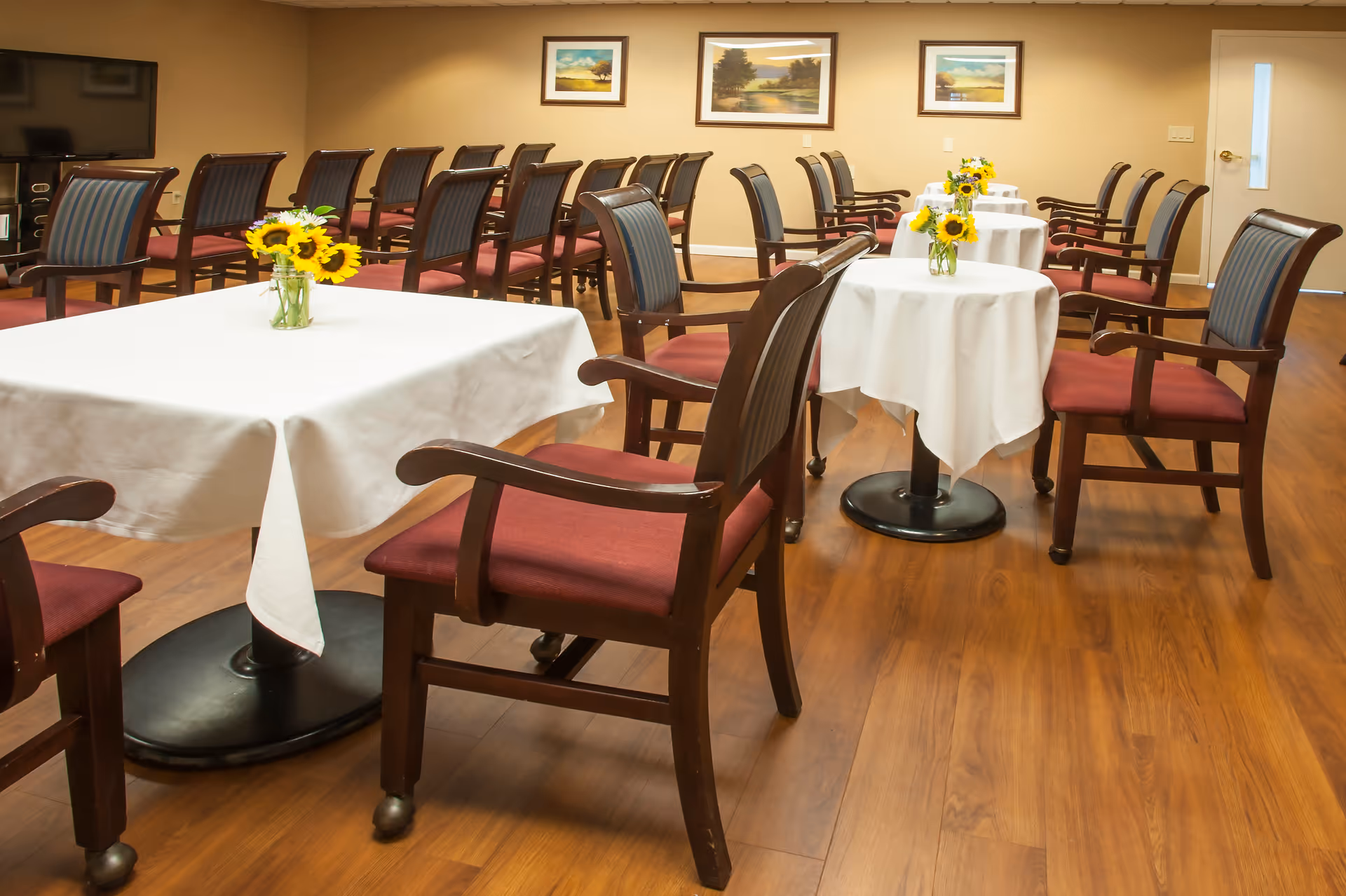 A dining area with several tables covered with white tablecloths, each adorned with a small vase of sunflowers. Wooden chairs with red cushions are arranged around the tables and in rows along the wall. The room has wooden flooring and beige walls decorated with framed landscape paintings.
