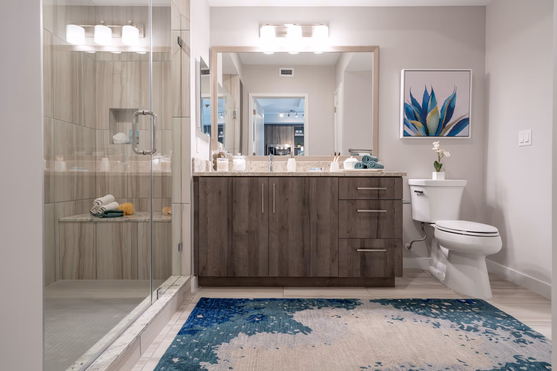 A modern bathroom featuring a glass-enclosed shower with beige tiles, a wooden vanity with a large mirror and countertop accessories, a toilet with a small potted plant on top, a framed blue and green plant artwork on the wall, and a blue and beige patterned rug on the floor.
