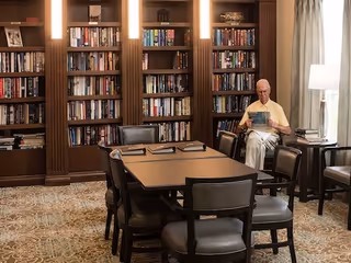 A senior man sitting on a chair reading a newspaper in a cozy library room with wooden bookshelves filled with books, a table with chairs in the center, and a lamp on a side table near a window with curtains.
