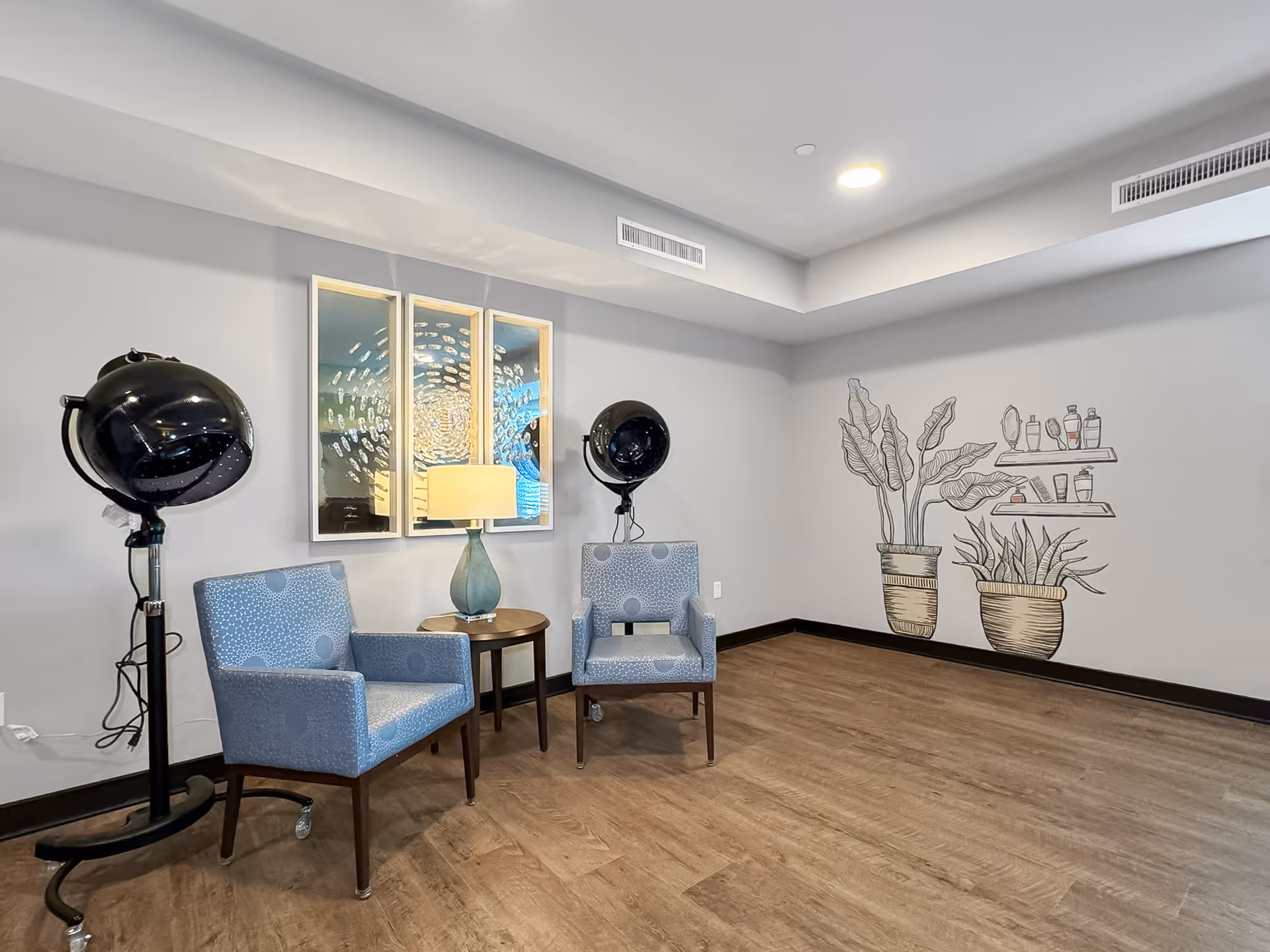 A small salon area with two blue patterned chairs and a round wooden table with a lamp between them. There are two black hair dryer chairs on either side of the chairs. On the wall behind, there are three framed mirrors and a wall mural depicting two potted plants and shelves with hair care products. The floor is wooden and the walls are light gray.