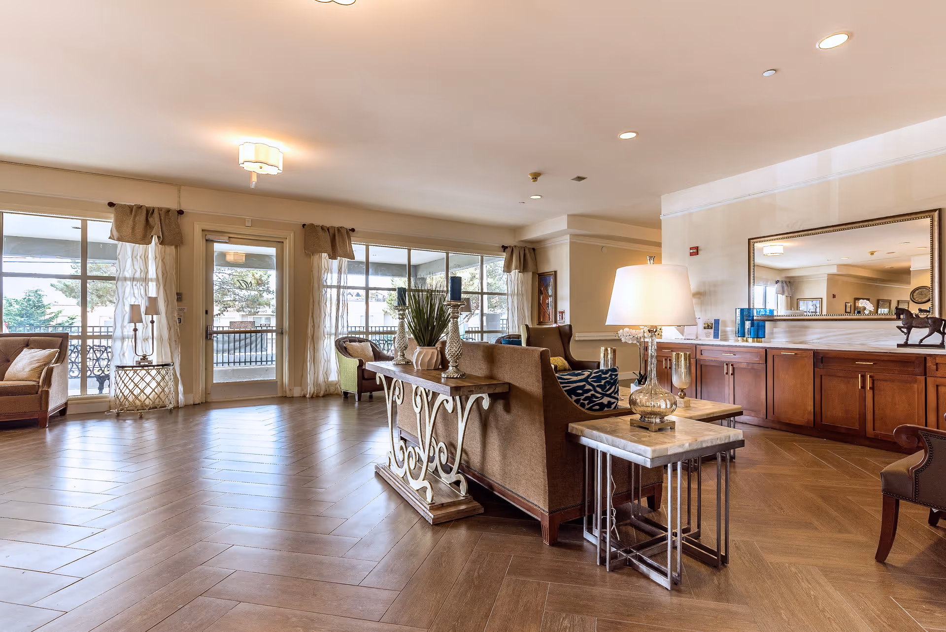 A spacious and well-lit senior living facility common area with wooden flooring, comfortable armchairs, a sofa, side tables with lamps, decorative plants, and large windows with sheer curtains allowing natural light to fill the room.